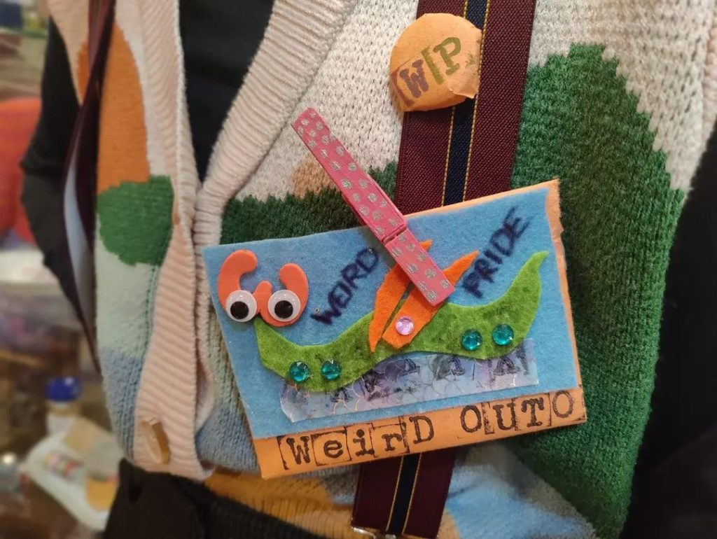 A Weird Pride badge made with felt, googly eyes, little jewel things and other adornments. 

At the bottom it seems to say WEIRD OUTO.